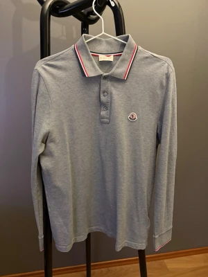 Grå långärmad skjorta från Moncler - Grå långärmad skjorta från Moncler med klassisk krage, knappar framtill och Moncler-logga på bröstet. Snygga röda och vita detaljer på krage och ärmslut ger en sportig touch. Tillverkad i mjuk bomull för skön känsla.