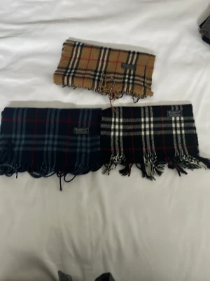 Burberry rutiga halsdukar i ull - Tre klassiska Burberry halsdukar i ull med ikoniskt rutmönster. En är beige med svarta och röda rutor, en är mörkröd med grått och vitt, och en är mörkblå med röda och ljusblå rutor. Alla har fransar i ändarna och Burberry-logga.