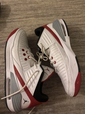 Vita och röda Air Jordan sneakers - Säljer ett par vita Air Jordan sneakers med röda och grå detaljer. Skorna har perforerat läder, svarta insidor och klassisk Jumpman-logga på plösen. Snygg design med lågt skaft och bekväm sula. Perfekta för dig som gillar streetwear och sportig stil.