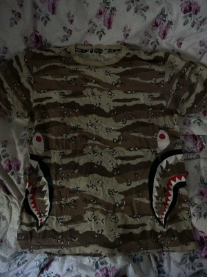 Bape  - Cool autentisk t-shirt från bape med klassiskt camouflage-mönster i beige och bruna toner. Tryck med hajmotiv på sidorna fram/bak. Perfekt för dig som vill sticka ut med streetwear-stil.