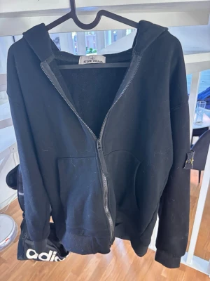 Svart hoodie från Stone Island - Säljer en svart hoodie från Stone Island med dragkedja framtill och klassisk logotyp-patch på vänster ärm. Hoodien har huva, två fickor och ribbade muddar. Tillverkad i mjukt bomullsmaterial som är skönt att bära.