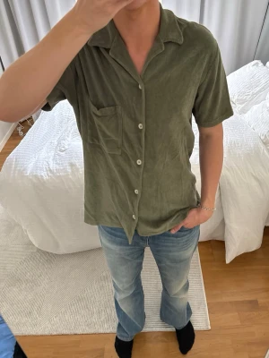 Olivgrön kortärmad skjorta i frotté - Säljer en olivgrön skjorta i mjuk frotté med korta ärmar, klassisk krage och bröstficka. Skjortan har vita knappar framtill och en avslappnad passform som ger en chill vibe.
