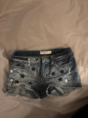 Blå jeansshorts med stjärnor från Lindex - Snygga blå jeansshorts från Lindex med slitna detaljer och mörka stjärnor broderade på framsidan. Klassisk femficksmodell med råa kanter och knappgylf. Perfekta för en avslappnad och trendig look.