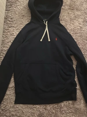 Svart hoodie från Polo Ralph Lauren - Klassisk svart hoodie från Polo Ralph Lauren med vit dragsko i huvan och röd broderad logga på bröstet. Hoodien har känguruficka framtill och är gjord i mjukt bomullsmaterial. Perfekt för en chill och stilren look.
