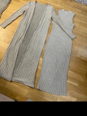 Grå ribbat set med lång kofta - Säljer ett snyggt tvådelat set i ljusgrå ribbstickad trikå. Setet består av en ärmlös lång bodyklänning och en matchande lång kofta med långa ärmar. Perfekt för en trendig och bekväm look. Materialet är mjukt och stretchigt för extra komfort.