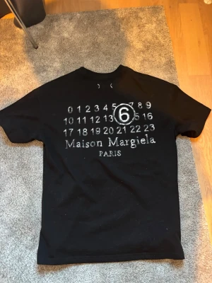Maison Margiela svart t-shirt - Svart t-shirt från Maison Margiela med ikoniskt tryck av siffror och stor vit 6:a på bröstet. Klassisk passform och rund halsringning. Perfekt för dig som gillar streetwear och designerplagg.