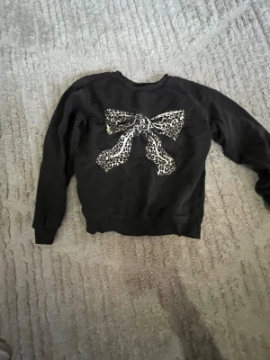 Svart hoodie med leopardrosett - Cool svart hoodie med stor rosett framtill i leopardmönster. Perfekt för dig som vill sticka ut lite extra. Mjuk och skön med klassisk passform och rund hals.