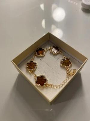 Guldarmband med bruna blommor - Snyggt armband i guldton med fem fyrklöverformade hängen i brun sten och glittrande kanter. Justerbar kedja med karbinlås. Perfekt accessoar för att lyfta din outfit med en unik och trendig touch.