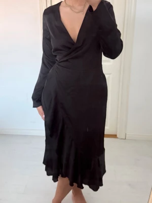 Svart omlottklänning med volang - Svart långklänning med omlottmodell och djup v-ringning. Klänningen har långa ärmar och volangdetalj nertill, vilket ger en snygg och trendig siluett. Perfekt för dig som gillar stilrena och eleganta plagg.