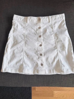 Vit jeans kjol med knappar framtill - Snygg vit jeanskjol med knappar längs hela framsidan. Kjolen har en rak passform och markerade sömmar som ger en stilren look. Perfekt att matcha med en crop top eller en oversized hoodie. Står storlek L i plagget men skulle mer säga att det är storlek M. 