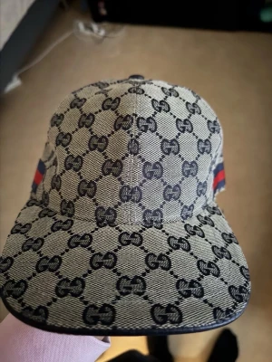Snygg keps med fint G mönster - Snygg keps med klassiskt monogram i svart på beige bakgrund. Kepsen har böjd skärm och röd-blå rand på sidan. Tillverkad i bomullsmaterial och perfekt för dig som vill sticka ut med en ikonisk accessoar.