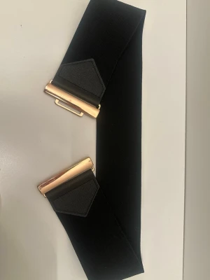 Svart brett midjebälte med gulddetalj - Snyggt svart midjebälte med bred resår och guldfärgat spänne framtill. Perfekt för att ge extra form till klänningar eller oversized skjortor. Bältet har en enkel och stilren design utan mönster.