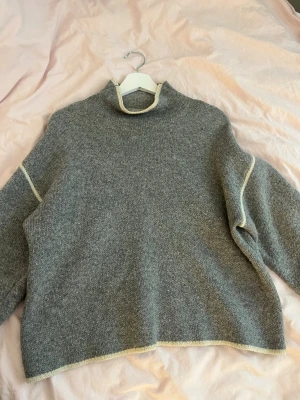 Grå stickad tröja från Zara - Mysig grå stickad tröja från Zara med ljusa kontrastsömmar vid hals, ärmar och nederkant. Tröjan har en lösare passform och polokrage. Säljer då den inte längre används!