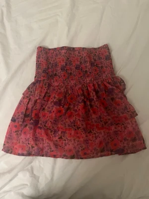 Blommig volangkjol från BikBok - Supersöt kort kjol från BikBok med blommigt mönster i rosa och röda toner. Kjolen har smock i midjan och volanger som ger en flowy känsla. Perfekt för dig som gillar färg och vill sticka ut lite extra. Den är endast använd 1 gång så i ett väldigt bra skick❤️❤️