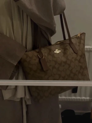 Brun axelväska från Coach - Snygg brun axelväska från Coach med klassiskt monogrammönster i canvas och detaljer i läder. Väskan har två långa axelband, dragkedja upptill och en hängande tag med Coach-logga. Perfekt för att bära allt du behöver med stil. Liten defekt som knappt syns