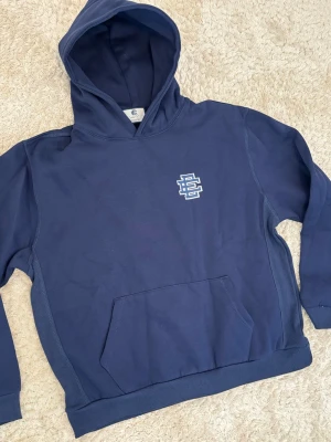 Mörkblå hoodie  - Snygg mörkblå hoodie från med vit logga på bröstet. Klassisk känguruficka framtill och stor huva. Tillverkad i mjuk bomull för skön känsla. Perfekt för dig som gillar streetwear och vill ha en stilren look.