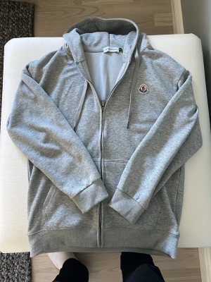 Moncler zip tröja  - Snygg grå zip tröja från Moncler i storlek M som är äkta, använd 1-2 gånger men passar inte mig.  Tröjan har huva med dragsko och ribbade muddar vid ärmar och nederkant. Tillverkad i mjuk bomullsblandning som känns skön mot huden. Perfekt för en avslappnad och trendig look. Påse medföljer! Hör av er om ni har frågor eller vill ha mått!