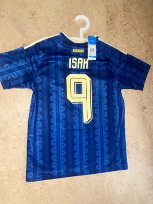 Blå Sverige VM fotbollströja Isak 9 - Säljer en blå Sverige fotbollströja från Adidas med Isak och nummer 9 på ryggen. Tröjan har gula detaljer, korta ärmar och ett mönster i olika blå nyanser. Perfekt för dig som vill stötta landslaget med stil.