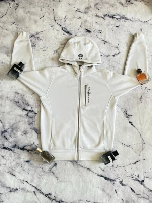 Vit hoodie från Sail Racing - Vit hoodie med dragkedja från Sail Racing. Har broderad logga på bröstet och stor text 'SAIL RACING EST. 1977' på ryggen. Utrustad med huva och två fickor framtill. Snygg och stilren design, perfekt för en sportig look.