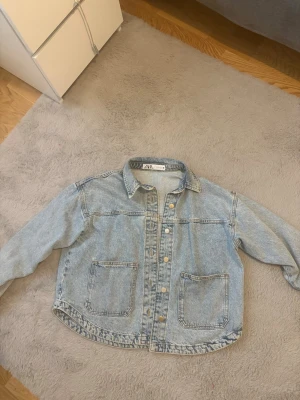 Ljusblå jeansjacka från Zara - Snygg ljusblå jeansjacka från Zara med klassisk krage, två stora fickor framtill och knäppning med metallknappar. Jackan har en avslappnad passform och är perfekt för en trendig vardagslook.