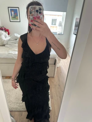 Svart långklänning med volanger - Elegant svart långklänning med volangdetaljer och v-ringning. Klänningen har ärmlös design och öppen rygg med tunna axelband. Inköptes nyligen till min Studentskiva och har endast använts en gång. Den har en dragkedja på sidan. Och det går att justera banden i ryggen💞