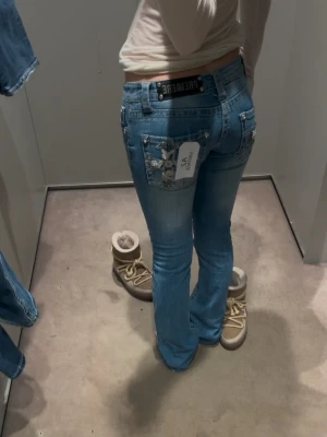 Ljusblå jeans från Rue21 - Säljer ett par ljusblå jeans från Premage med utsvängda ben och snygga detaljerade bakfickor med stjärnor och paljetter. Jeansen har låg midja och klassisk femficksdesign. Perfekta för dig som vill sticka ut med coola detaljer! Passar som W25 L30/32. Midjemått rakt över 26, innerbenslängd 78