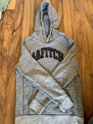 Grå hoodie Abercrombie & Fitch - Säljer en grå hoodie från Abercrombie & Fitch med stor svart logga på bröstet. Hoodien har huva med dragsko och en mjuk, fleecefodrad insida. Klassisk känguruficka framtill och ribbade muddar vid ärmar och nederkant. Perfekt för chill dagar.