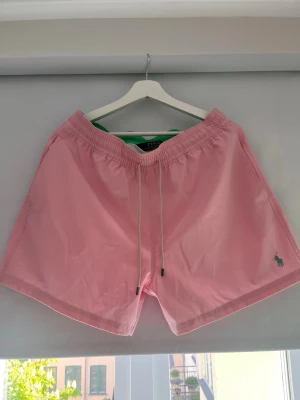 Rosa badbyxor från Polo Ralph Lauren - Säljer ett par ljusrosa badbyxor från Polo Ralph Lauren med elastisk midja och vita dragsnören. Klassisk passform, liten grön logga broderad framtill och ficka med knapp baktill. Perfekta för stranden eller poolen.