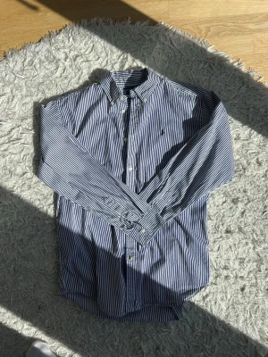 Ralph Lauren Randig blåvit skjorta - Klassisk skjorta från Ralph Lauren i blåvitt randigt mönster. Skjortan har lång ärm, knappar framtill och en liten broderad logga på bröstet. Perfekt för en stilren look och tillverkad i bomull för skön känsla. Står XL men sitter mer som en medium, skickar gärna mått om du är osäker på hur den passar! Hör gärna av er vid frågor. Mvh Isak