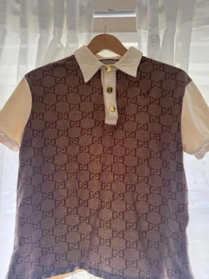 GUCCI GG-Jacquard Polo Shirt - Gucci topp med korta ärmar, vit krage och knappslå framtill. Framsidan har ett brunt GG-mönster och ärmarna är beige. Tidlös design med tre guldiga knappar och klassisk passform. Perfekt för dig som vill sticka ut med lyxig känsla.