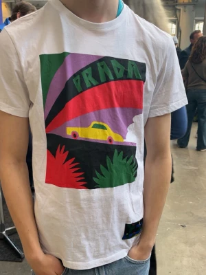 Prada ”graphic printed tshirt” - Vit t-shirt från Prada med stor färgglad grafisk print framtill som visar en gul bil och färgstarka former. Modellen på bilden är storlek S, tröjan passar dig som är S och mindre. En tröja som är väldigt svår att få tag på. (Se sista bild). Prada har sortiment i samma grafisk design på flera olika tröjor.