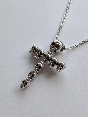 Silverfärgat halsband med dödskallekors - Unikt halsband med ett korsformat hänge av flera små dödskallar i blankt silverfärgat metall. Kedjan är tunn och matchar hängets coola och edgy stil. Perfekt accessoar för dig som gillar alternativt mode och vill sticka ut. Materialet rostfritt stål. 