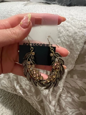 Guldiga örhängen med strass och fjäderform - Snygga örhängen i guldig metall med fjäderformad design och glittrande strass-stenar. Perfekta för att ge din look lite extra bling! Hängande modell med krok, passar dig som gillar statement-smycken.