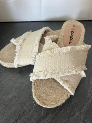 Beiga sandaler med fransade band Bonprix - Säljer ett par beiga sandaler från Bonprix med breda korslagda band i tyg och fransade kanter. Sulan är i flätad jute-look och undersidan har gummisula. Perfekta för en avslappnad och trendig sommarstil.