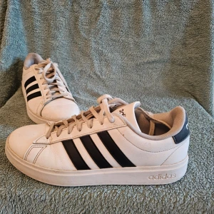Vita Adidas sneakers med svarta ränder - Klassiska vita sneakers från Adidas med de ikoniska tre svarta ränderna på sidorna och svart detalj vid hälen. Skorna har lågt skaft, snörning och är tillverkade i syntetmaterial med textilfoder. Perfekta för en sportig och stilren look.