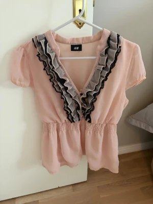 Ljusrosa/beige volangblus från H&M - Supersöt ljusrosa/beige blus från H&M med korta puffärmar och djup v-ringning. Framtill har den dubbla volangdetaljer i svart och grått som ger en romantisk vibe. Köpt på plick men passar inte mig tyvvär 😭 står ingen storlek men skulle säga M men passar även S
