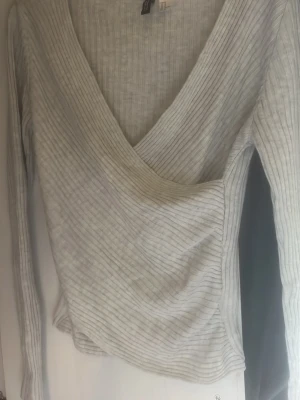 Ljusgrå omlottopp från H&M - Säljer en ljusgrå ribbad omlottopp från H&M med lång ärm och v-ringad hals. Toppen har en smickrande omlott-design och är tillverkad i ett mjukt, stretchigt material som sitter snyggt på kroppen.