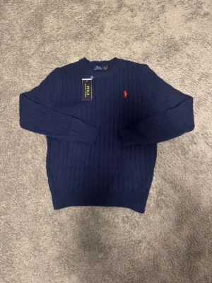 Mörkblå kabelstickad tröja Polo Ralph Lauren - Mörkblå kabelstickad tröja från Polo Ralph Lauren med rund halsringning och den klassiska röda logotypen broderad på bröstet. Tröjan har långa ärmar och ribbade muddar vid ärmslut och nederkant. Perfekt för dig som gillar stilren och tidlös design.
