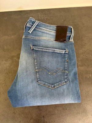 Ljusblå raka jeans från Replay 33/34 - Snygga ljusblå jeans från Replay med raka ben och klassisk femficksmodell. Jeansen har slitna detaljer och en cool kontrastsöm på bakfickan samt ett brunt läderpatch i midjan. Perfekta för en avslappnad och trendig look.