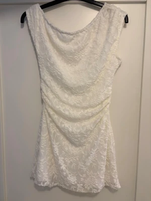 Vit spetsklänning med drapering - Säljer en vit kort klänning med vackert mönster och draperad effekt. Klänningen är ena sidan offshoulder men är gjord att man kan ha den på båda sidorna och har en elegant passform. Perfekt för sommarens alla tillfällen.