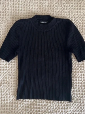 Svart ribbad topp från Gina Tricot - En svart ribbad topp från Gina Tricot med korta ärmar och högre rund halsringning. Toppen har en slim passform och är tillverkad i ett stretchigt material som sitter snyggt på kroppen. Perfekt att styla med jeans eller kjol.