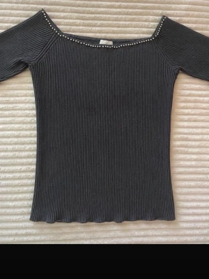 Grå ribbad offshoulder topp - Trendig svart ribbad topp med offshoulder-modell och små silverfärgade pärlor längs halsringningen. Figurnära passform och stretchigt material som sitter snyggt på kroppen. Perfekt för dig som gillar stilrena detaljer.