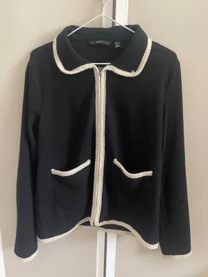 Svart cardiganjacka med vita detaljer - Stilren svart cardiganjacka från H&M med vita dekorativa kanter längs krage, dragkedja, fickor och ärmslut. Jackan har två fickor framtill och är tillverkad i ett mjukt tyg som ger en klassisk look. Perfekt för dig som gillar en chic och enkel stil.