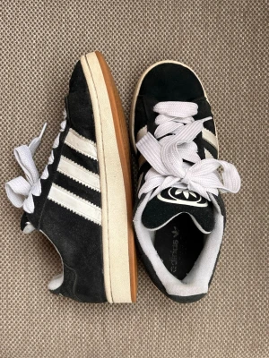 Adidas Campus svarta sneakers - Svarta Adidas Campus sneakers i mocka med vita detaljer och klassiska tre ränder på sidorna. Skorna har vit sula, vita skosnören och Adidas-logga på plösen och hälen. Låg modell med retrovibe och bekväm passform.
