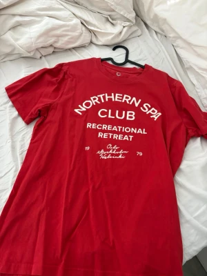 Röd t-shirt Northern Spa Club - Röd t-shirt med vit text 'Northern Spa Club Recreational Retreat' framtill. Klassisk rund halsringning och korta ärmar. 