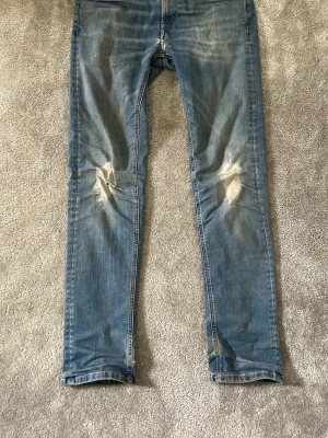 Blå slitna jeans från Replay - Säljer ett par blå jeans från Replay med slitningar och hål på knäna. Jeansen har klassisk femficksdesign, normal passform och är tillverkade i denim. Perfekta för en avslappnad och trendig look.