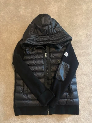 Svart pufferjacka med huva från Moncler - Snygg svart cardigan från Moncler med stickade ärmar och vadderad kropp. Jackan har huva, dragkedja framtill och en Moncler-logga på ärmen. Perfekt nu till våren. Har NFC tagg samt scan inuti.