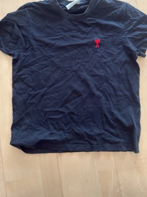 Svart t-shirt från Ami Paris - Snygg svart t-shirt från Ami Paris med klassisk passform och korta ärmar. På bröstet sitter Ami:s ikoniska röda hjärta-broderi. Tillverkad i mjuk bomull som känns skön mot huden. Perfekt till jeans eller shorts för en clean look.
