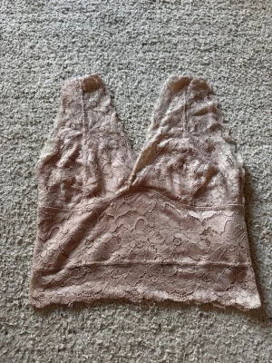 Ljusrosa spetsbody med V-ringning - Säljer en ljusrosa/beige topp i mjuk spets med djup V-ringning och breda axelband. Perfekt att styla under en skjorta eller kavaj.