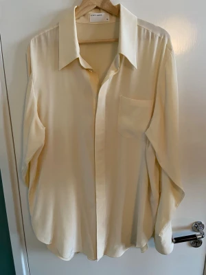 Klassisk butter yellow silkesskjorta från A Days March - Stilren butter yellow skjorta från Days March i silke material. Skjortan har klassisk krage, bröstficka och knäppning framtill. Passformen är loose och ärmarna är långa med knappar vid manschetterna. Oversize-stil